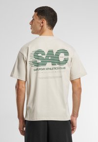 Béžové bavlněné tričko s velkým zeleným potiskem loga "SAC" na zádech, s horizontálními linkami a textem pod ním. Volný střih, krátké rukávy.
