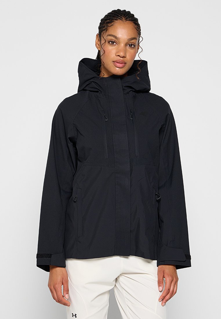 Kobieta w czarnej wodoodpornej kurtce z kapturem, z zamkiem i mankietami na rzep, w połączeniu z białymi spodniami Under Armour, stojąca na jednolitym tle.