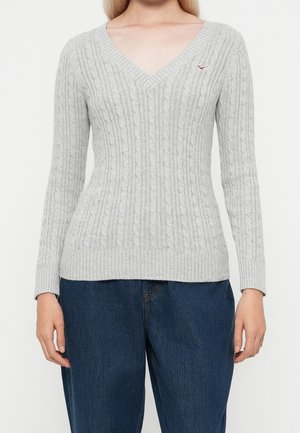 Mujer con un suéter de punto grueso color gris claro con cuello en V y jeans azul oscuro, de pie frente a un fondo blanco liso.