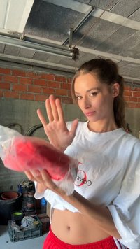 Mulher com um top curto branco e calções vermelhos segurando um objeto vermelho embrulhado com uma mão levantada, de pé num armazém com paredes de betão e prateleiras.