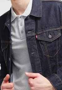 Giacca di denim scuro con cuciture dorate, dotata di colletto a punta, tasche frontali e bottoni, indossata sopra una polo grigia chiara.