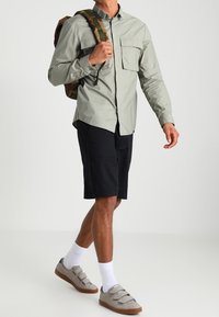 Mężczyzna ubrany w jasnoszary długi rękaw, czarne szorty, białe skarpetki, szare sneakersy i noszący plecak w kamuflażu na ramieniu.