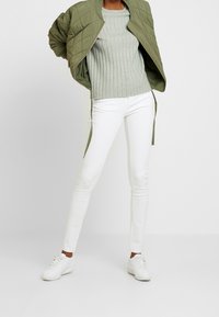 Vadderad olivgrön bomberjacka, ljusgrå ribbad tröja, vita slim-fit jeans och vita sneakers. Enkel och avslappnad outfit.