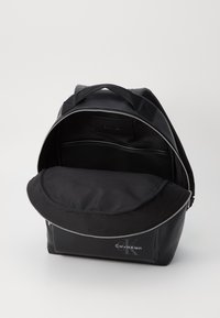Črn nahrbtnik iz gladkega sintetičnega materiala, z zadrgo, velikim zunanjim žepom in nastavljivimi naramnicami. Na sprednji strani ima logo Calvin Klein.
