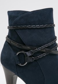 Botín de ante azul marino con tacón alto, que presenta correas de tejido negro y un anillo decorativo, mostrando un diseño elegante y afilado.