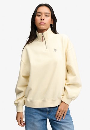 Ung kvinde iført cremefarvet lynlåstrøje og blå jeans, stående med den ene hånd hvilende på hoften.