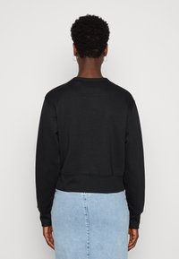 Personne avec des cheveux bouclés courts et des boucles d'oreilles en forme de créole, portant un haut à manches longues noir et une jupe en denim bleu clair, de dos contre un fond uni.