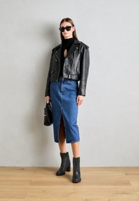 Giubbotto da motociclista corto in pelle nera sopra un top nero, gonna midi in denim con uno spacco frontale e stivali neri con tacco e lacci. Occhiali da sole neri.