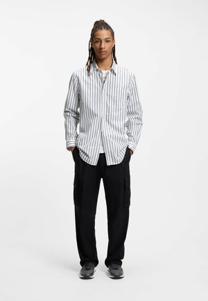 Striped button-up shirt in white and navy, paired with black cargo pants. Footwear is gray sneakers. Model stands in a neutral background.