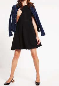 Svart klänning med en utsvängd kjol och textilt tyg, i kombination med en blå jeansjacka. Modellen bär svarta pumps med spetsig tå.