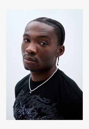 Camiseta gráfica negra con diseño blanco, acompañada de un collar de cuentas plateadas. El sujeto tiene el cabello corto trenzado y una expresión neutral.