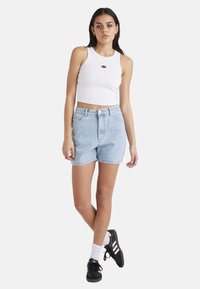 Weißes geripptes Tanktop mit kleinem schwarzem Logo, kombiniert mit hellblauen, hochgeschnittenen Denimshorts. Das Model trägt schwarze Sneakers mit weißen Socken.