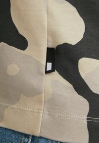 Camiseta de camuflaje con patrones abstractos en beige y negro, fabricada en un tejido suave. Presenta un dobladillo cosido y una pequeña etiqueta negra en el lateral.