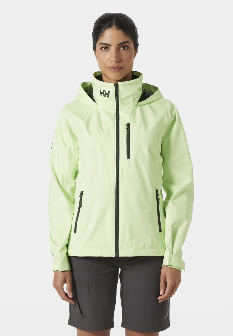 Chaqueta impermeable de color verde claro con capucha, con cremalleras y acentos negros, y un bolsillo en el pecho con cremallera. La lleva puesta con pantalones cortos oscuros.
