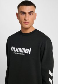 Ung man med kort hår som bär en svart sweatshirt med "hummel SPORTSWEAR SINCE 1923" och vita chevronmönster på ärmarna.