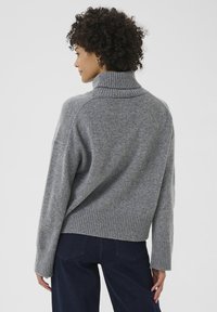 Grauer Strickpullover mit hohem Kragen, geripptem Saum und Bündchen, verkürztem Rückenteil und weicher Textur. Getragen mit dunkelblauen Hosen.