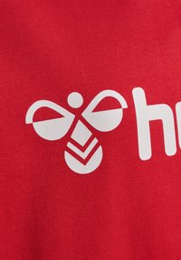 Roter Stoff mit weißem Grafikmotiv einer stilisierten Biene mit runden Flügeln und gestreiftem Körper, begleitet vom Text "he".