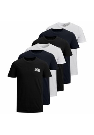Jeune homme ajustant son col, portant un t-shirt blanc Jack & Jones et un pantalon foncé sur un fond uni.