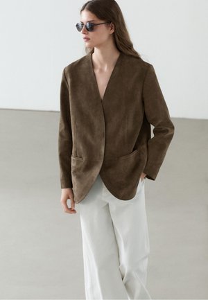 Frau trägt dunkelbraune Wildlederjacke und weiße Hosen mit weitem Bein, mit Sonnenbrille, stehend in minimalistischem Innenraum.