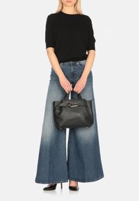 Borsa tote in pelle nera con finitura liscia, forma quadrata, manici corti e un accento con cinturino rettangolare. Indossata con pantaloni in denim a gamba larga.