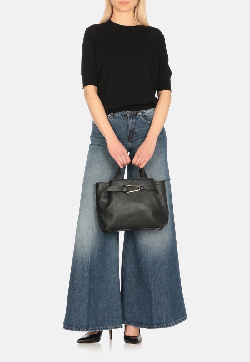 Borsa tote in pelle nera con finitura liscia, forma quadrata, manici corti e un accento con cinturino rettangolare. Indossata con pantaloni in denim a gamba larga.