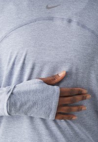 Světlý šedý atletický tričko s dlouhým rukávem a otvory na palce. Textura je měkká a pružná, s jemným melírovaným vzorem. Logo Nike je p�řítomné.