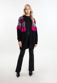 Svart cardigan med rosa och grå mönstrade detaljer, med fransade kanter. Bärs över en svart topp och matchas med svarta utsvängda byxor och stövlar.