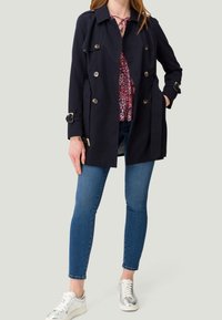 Granatowy płaszcz trencz z podwójnym zapięciem na guziki, złotymi guzikami, regulowanym paskiem w talii i luźnym krojem, noszony na bluzce w kwiatowy wzór i dżinsach.