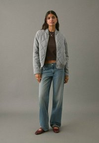 Mujer de pie contra un fondo gris, vistiendo una chaqueta gris acolchada, una parte superior oscura corta de color marrón, unos jeans anchos desgastados y unas bailarinas burdeos con tiras en el tobillo.