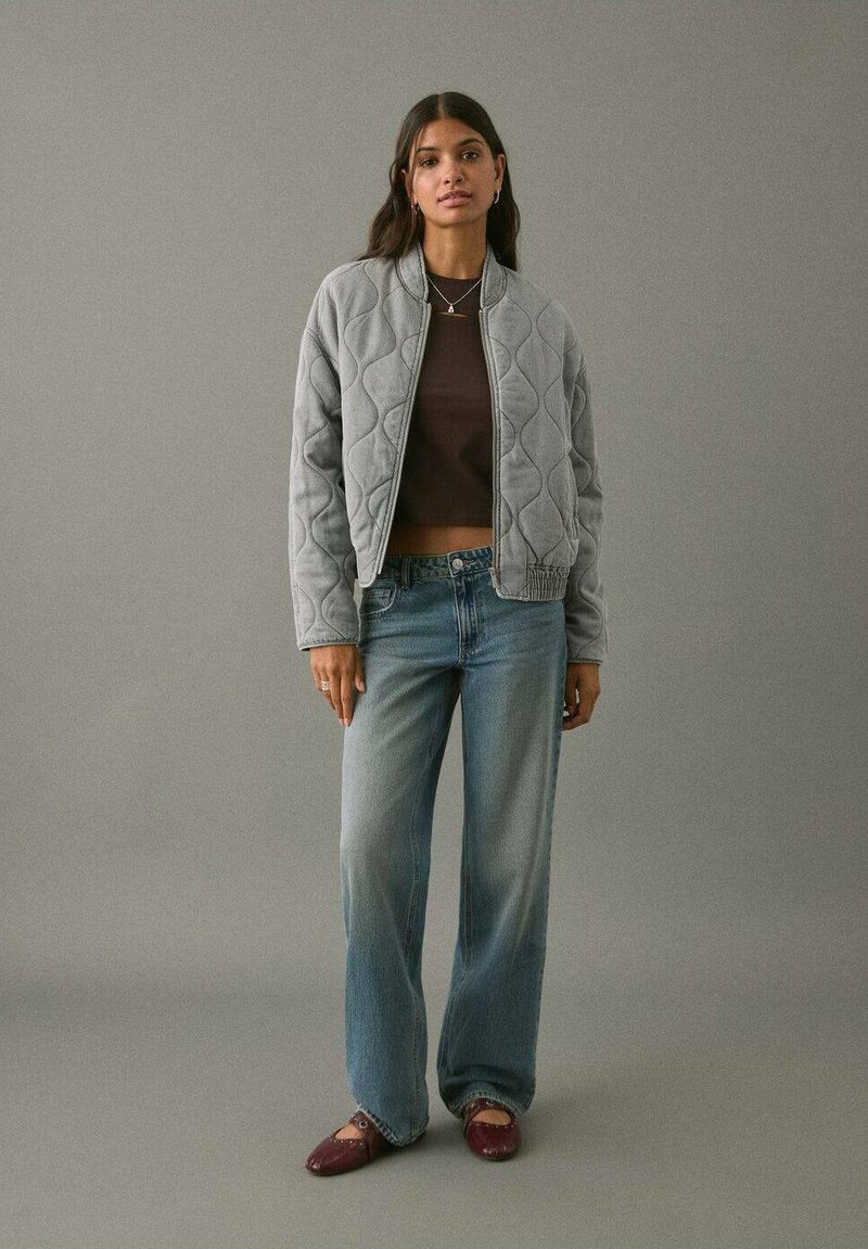 Mujer de pie contra un fondo gris, vistiendo una chaqueta gris acolchada, una parte superior oscura corta de color marrón, unos jeans anchos desgastados y unas bailarinas burdeos con tiras en el tobillo.