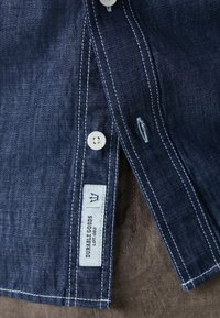 Camisa de mezclilla en azul oscuro con costuras blancas, con botones y una etiqueta de marca que dice "DURABLE GOODS". Primer plano de la tela texturada.