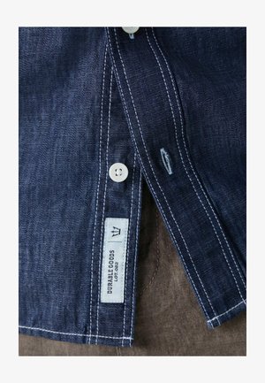 Tamna plava traper košulja s bijelim šavovima, s gumbima i brendiranim etiketom s natpisom "DURABLE GOODS". Blizu prikazane teksturirane tkanine.