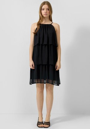 Młoda kobieta w czarnej sukience bez rękawów z falbanami i cienkimi ramiączkami oraz w czarnych siatkowych sandałach na obcasie stoi na jednolitym tle.