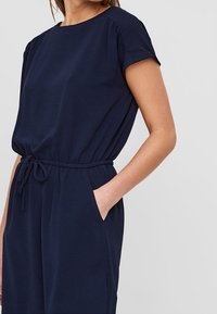 Námořnicky modrý overal s krátkým rukávem, staženým pasem a bočními kapsami. Vyroben z měkké tkaniny s uvolněným střihem a kulatým výstřihem.