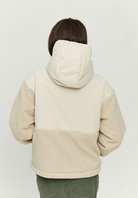 Beige hooded jacket with a textured fleece lower section and smooth upper fabric. Features a boxy design and a lined, quilted hood.