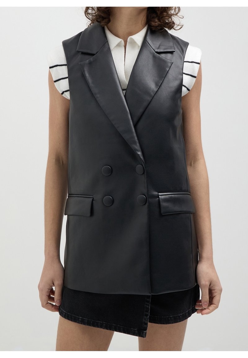 Svart faux läder ärmlös väst, dubbelknäppt design, med stora slag och sidofickor, bärs över en vit randig topp.