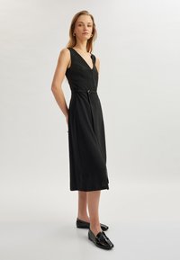 Zwarte midi-jurk met een v-hals, getailleerde taille en een zilveren ringaccent. Gemaakt van gladde stof, met brede banden en een vloeiende rok.