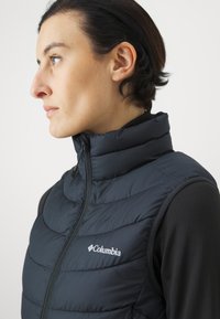 Crni prsluk od puffer materijala s visokim ovratnikom, s prednjim zatvaračem, kviltiranim dizajnom i Columbia logotipom s lijeve strane.