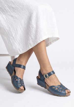 Pieds portant des sandales compensées en cuir bleu avec des motifs découpés et des brides à la cheville, assorties à une robe blanche longueur genoux.
