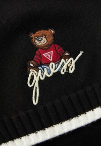 Svart stoff med en brodert brun bjørn som har p�å seg en rød skjorte med en trekanlogo og ordet "guess" i hvit skråskrift.