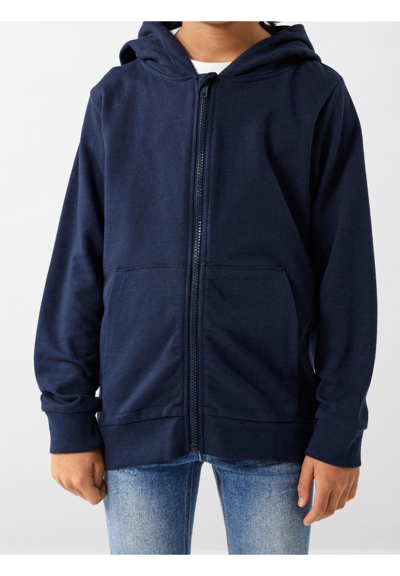 Felpa con zip di colore navy realizzata in un tessuto morbido, dotata di cappuccio, due tasche frontali e polsini a coste, indossata sopra un paio di jeans blu.