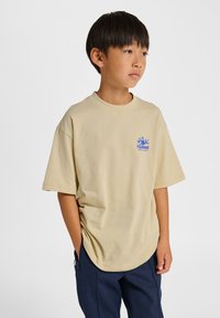 Beiges T-Shirt mit kurzen Ärmeln, rundem Ausschnitt und weiter Passform, mit einem blauen Logo auf der linken Brust. Kombiniert mit dunkelblauen Hosen.