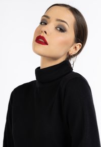 Zwarte coltrui met een zachte textuur. Het model heeft donker haar dat naar achteren is gestyled en draagt opvallende rode lippenstift, gecombineerd met grote creolen.