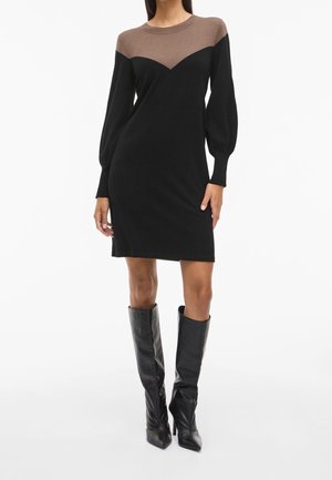 Femme portant une robe noire au genou avec un empiècement marron et des manches longues, associée à des bottes noires à talons montant jusqu'au genou.
