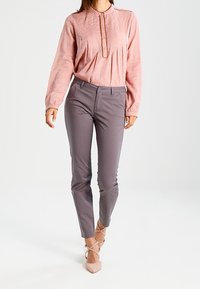 Blouse rose clair à manches longues avec col boutonné et texture subtile ; associée à un pantalon gris ajusté et des talons nus.
