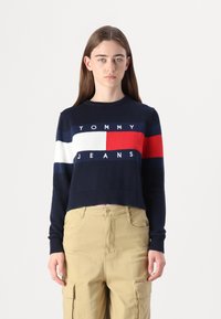Tamsiai mėlynas trumpas megztinis su balta, raudona ir mėlyna juostele, su "TOMMY JEANS" logotipu. Ribojo rankogaliai ir apačia, pagamintas iš megztos medžiagos.