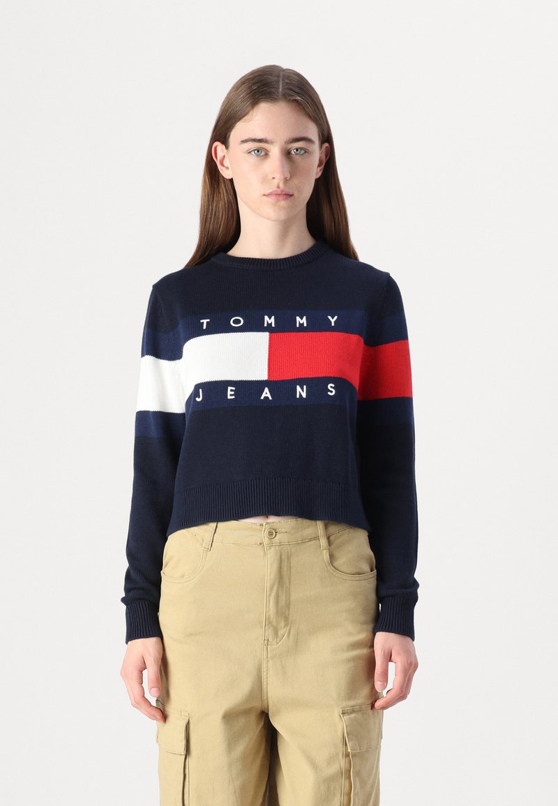 Tamsiai mėlynas trumpas megztinis su balta, raudona ir mėlyna juostele, su "TOMMY JEANS" logotipu. Ribojo rankogaliai ir apačia, pagamintas iš megztos medžiagos.