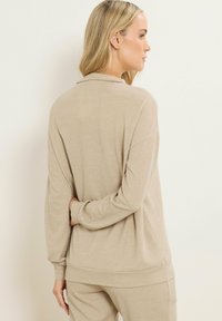 Donna con capelli biondi che indossa un top a maniche lunghe beigè a coste e pantaloni abbinati, in piedi con una mano dietro la schiena, rivolta verso destra.