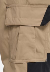 Μπεζ υφασμάτινη τσέπη με καπάκι και μαύρη λεπτομέρεια στη γωνία, σε μπεζ και μαύρο υφαντό φόντο.