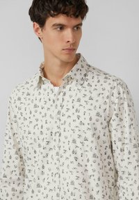 Biała koszula z delikatnym czarnym wzorem składającym się z różnych małych ilustrowanych obiektów. Posiada klasyczny kołnierzyk oraz zapinany na guziki przód.